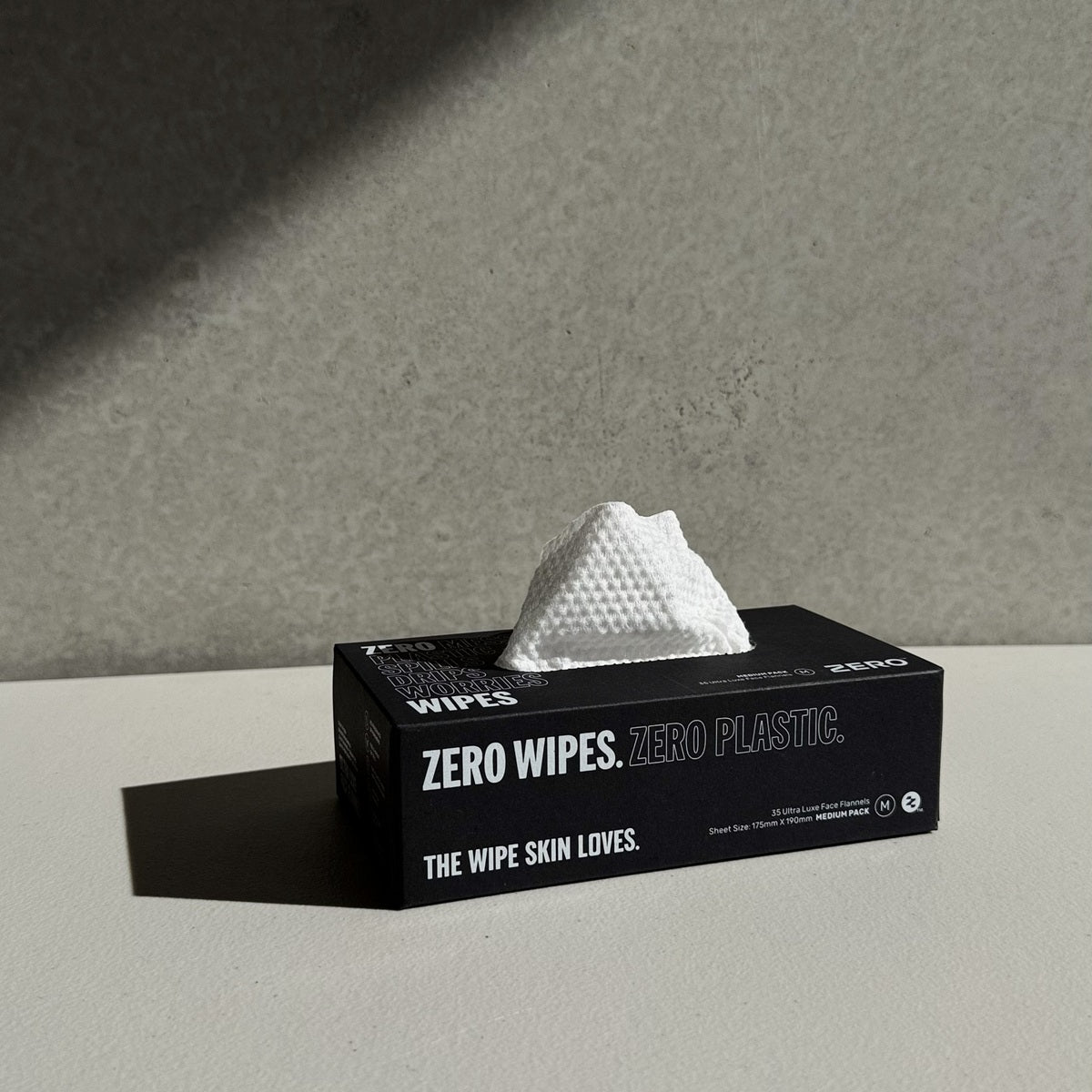 A black box of Zero Wipes tissue wipes sits on a light gray surface against a textured gray wall, with a white tissue peeking out from the top.