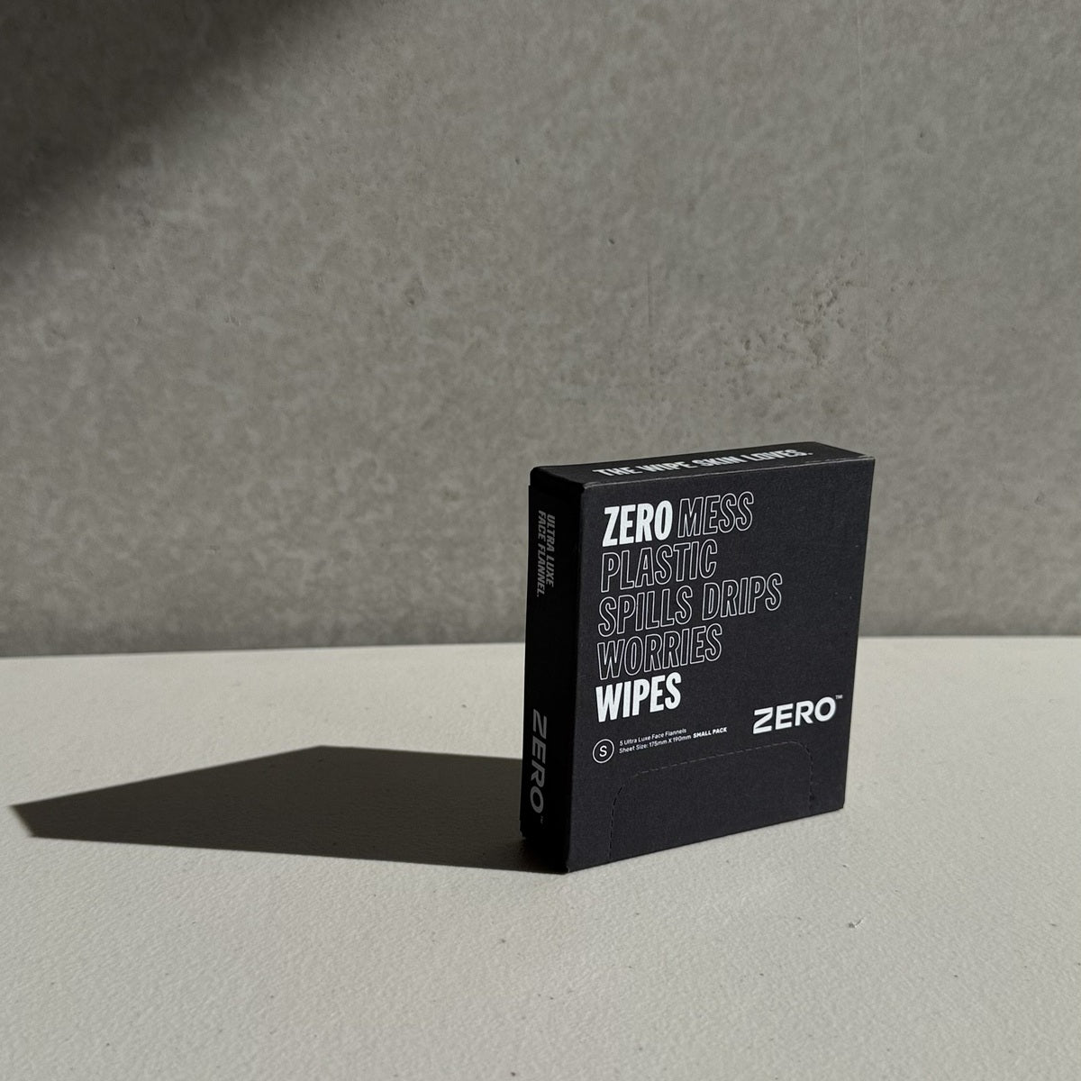 A black rectangular box of Zero Mess Plastics Spills Drips Worries Wipes standing upright on a light gray surface against a textured gray wall, casting a sharp shadow.
