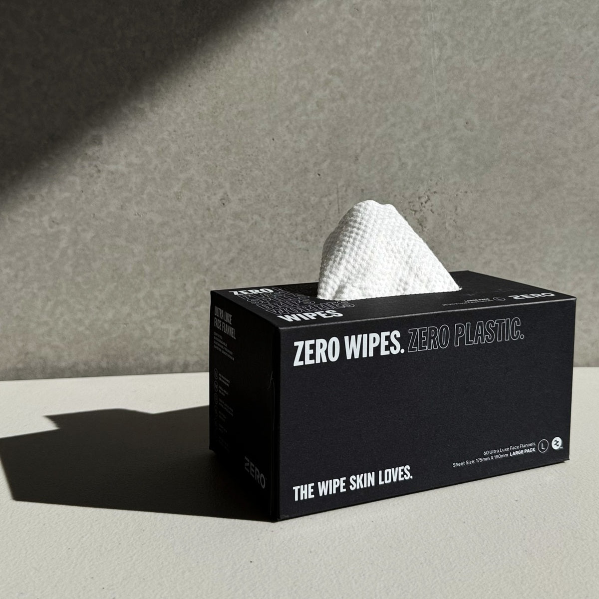 A black tissue box of zero waste facial wipes with a white textured wiper pulled from the top, set on a light gray surface against a textured gray background.