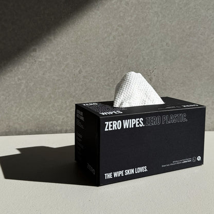 A black tissue box of zero waste facial wipes with a white textured wiper pulled from the top, set on a light gray surface against a textured gray background.